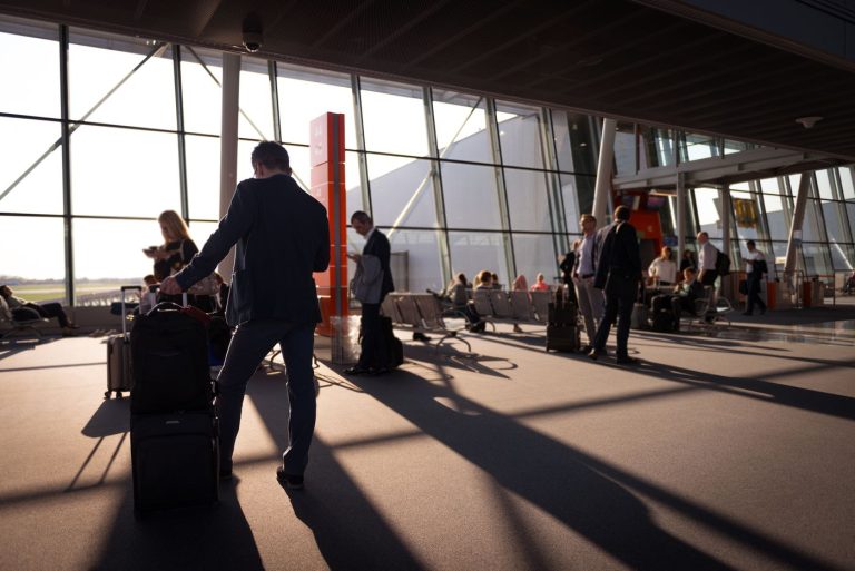 Passengers with luggage walking through a sunlit airport terminal