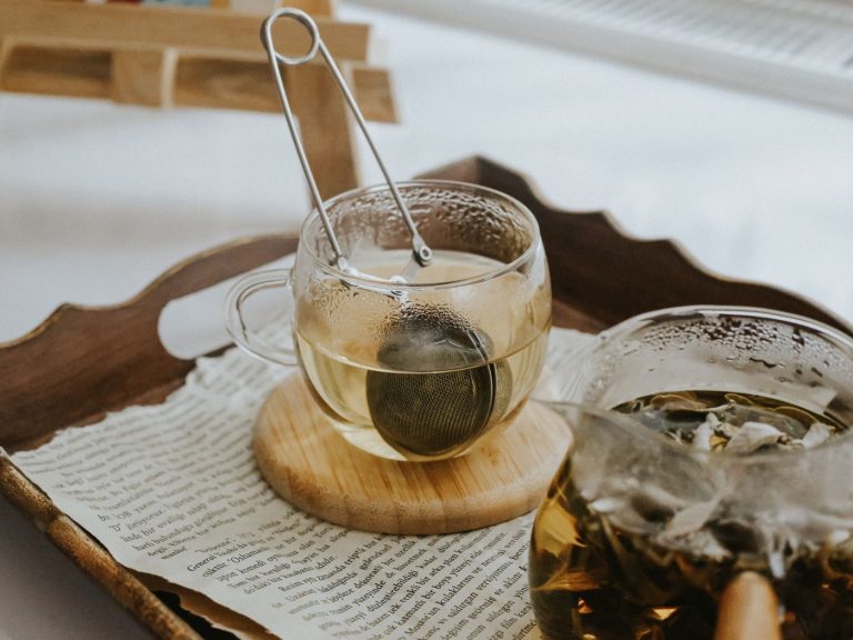 A glass of loose-leaf tea with a metal infuser resting on a wooden tray